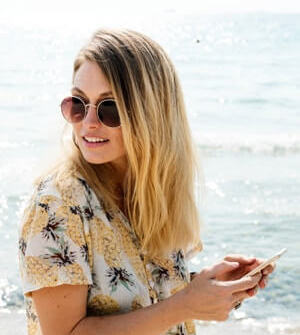 Blonde woman wearing sunglasses holding phone on beach